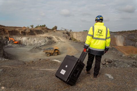Man wearing a yellow safety outfit and an Inspemon jacket, standing in front of a construction site and holding a suitcase.